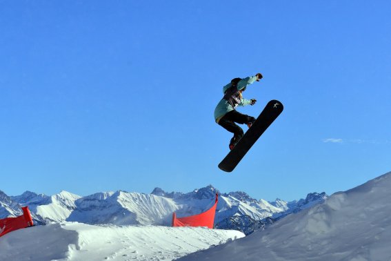 Ein Snowboardfahrer springt hoch über einen Schneehügel in einer winterlichen Berglandschaft. Es ist ein klarer, sonniger Tag.