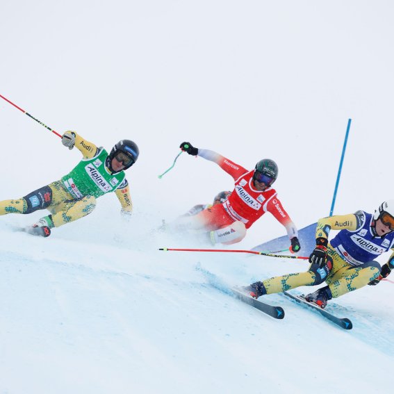Drei Skifahrer rasen bei einem Wettkampf in den Alpen auf einer verschneiten Piste.