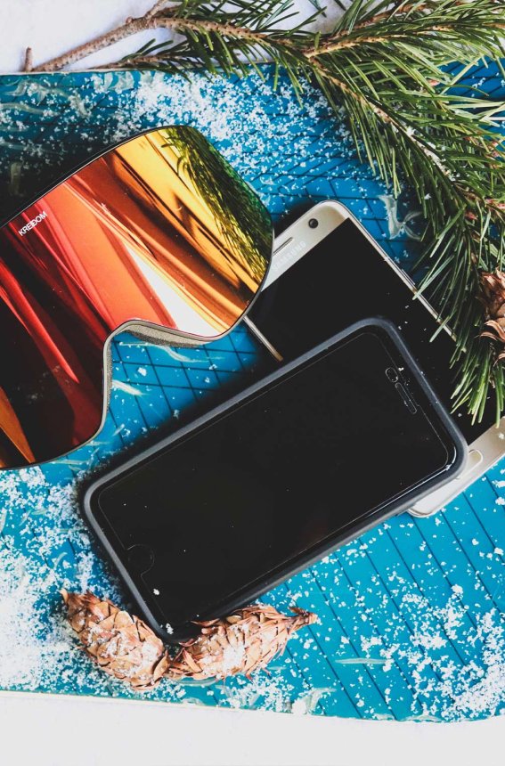 Auf einem Snowboard liegen eine farbige Skibrille, Handys und Tannenzweige. Perfekt für einen Skiurlaub im Winter.