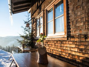 Die Berghütte steht im Winter, mit Tischen draußen und Blick auf die schneebedeckten Berge in der Ferne.