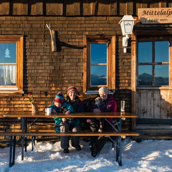 Zwei Freunde sitzen an einem Tisch vor einer gemütlichen Hütte, umgeben von Schnee und Bergen, und trinken warme Getränke.
