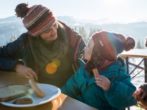 Eine Frau und ein Kind genießen ein herzhaftes Frühstück in den Bergen an einem sonnigen Wintertag.