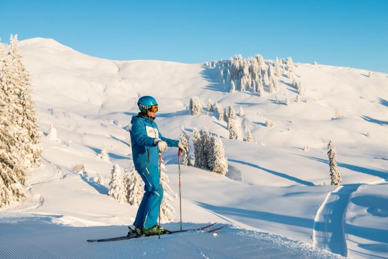 Ein Skifahrer steht auf der verschneiten Piste und genießt die Aussicht auf die schneebedeckten Berge und den blauen Himmel.