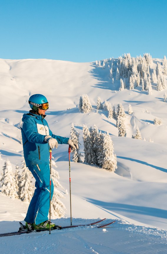 Ein Skifahrer steht auf der verschneiten Piste und genießt die Aussicht auf die schneebedeckten Berge und den blauen Himmel.