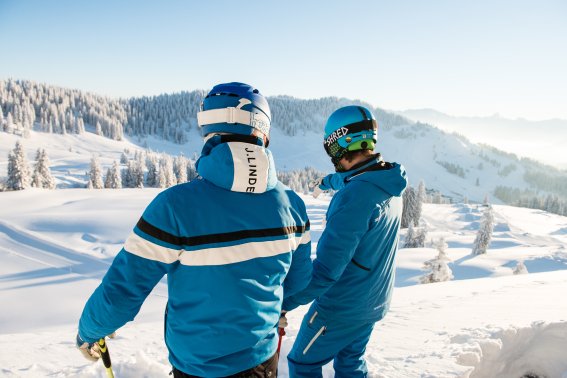 Ausblick auf die verschneite Berglandschaft im Winter Zwei Skifahrer stehen auf einem Aussichtspunkt und genießen die schöne winterliche Berglandschaft in der Ferne.