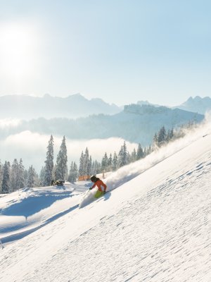 Zwei Wintersportler fahren den schneebedeckten Hang hinunter, umgeben von einem klaren blauen Himmel und schönen Bergen.