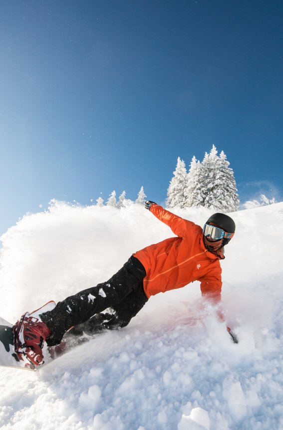 Ein Snowboarder in einer orangefarbenen Jacke fährt geschwind den Hang hinunter, umgeben von schneebedeckten Bäumen.
