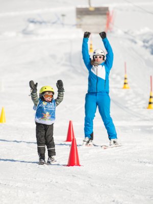 In einer schneebedeckten Landschaft üben Kinder das Skifahren mit einem Lehrer. Viele bunte Markierungen stehen herum.