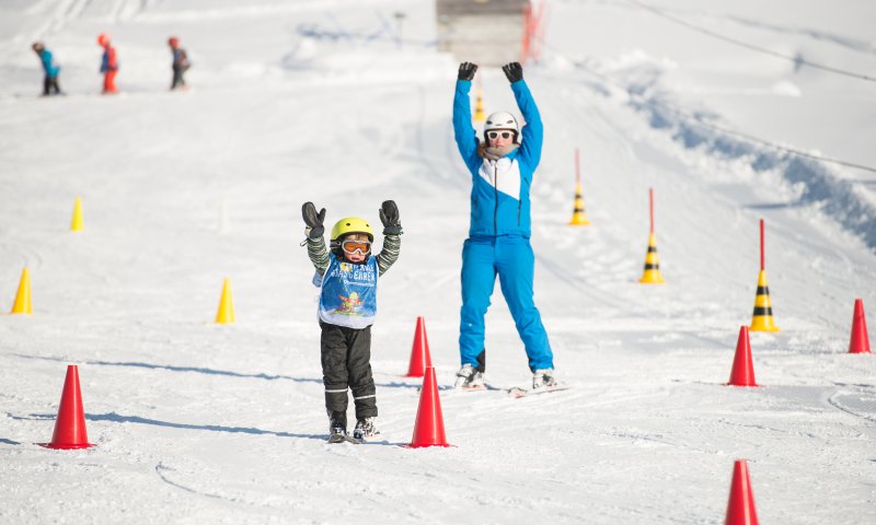 In einer schneebedeckten Landschaft üben Kinder das Skifahren mit einem Lehrer. Viele bunte Markierungen stehen herum.