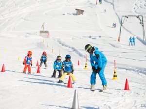 Eine Gruppe von Kindern übt das Skifahren mit einem Lehrer in einem schneebedeckten Skigebiet an einem sonnigen Wintertag.
