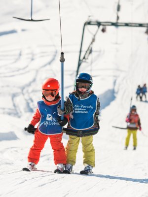 Zwei Kinder im Schnee warten auf den Schlepplift im Skigebiet. Der Himmel ist blau und viele Skifahrer sind in der Nähe.