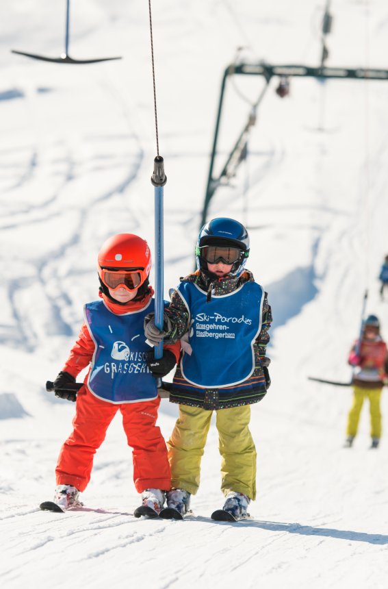 Zwei Kinder im Schnee warten auf den Schlepplift im Skigebiet. Der Himmel ist blau und viele Skifahrer sind in der Nähe.