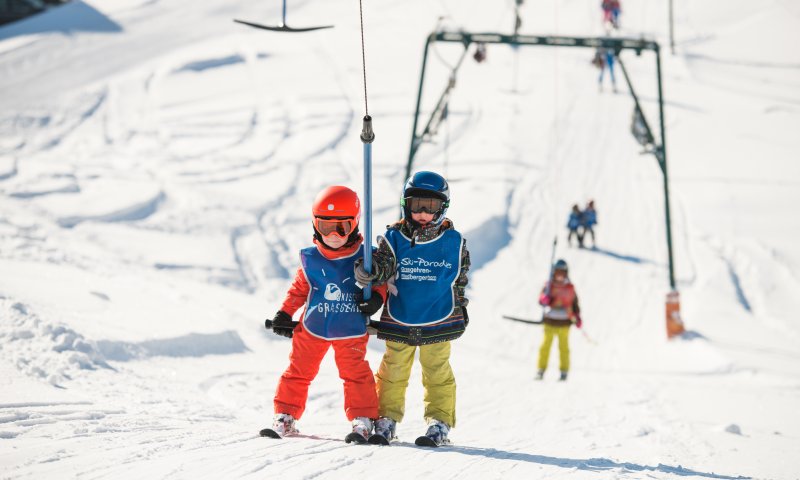 Zwei Kinder im Schnee warten auf den Schlepplift im Skigebiet. Der Himmel ist blau und viele Skifahrer sind in der Nähe.