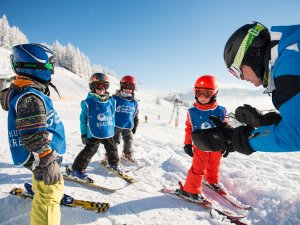 Vier Kinder üben das Skifahren mit einem Lehrer auf einer verschneiten Piste in den Alpen an einem sonnigen Wintertag.