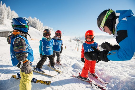 Vier Kinder üben das Skifahren mit einem Lehrer auf einer verschneiten Piste in den Alpen an einem sonnigen Wintertag.