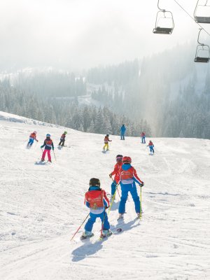 Kinder und Erwachsene fahren Ski auf den verschneiten Pisten eines Skigebiets in den Alpen an einem sonnigen Wintertag.