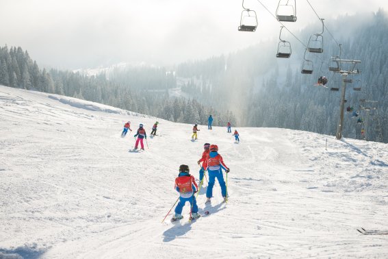 Kinder und Erwachsene fahren Ski auf den verschneiten Pisten eines Skigebiets in den Alpen an einem sonnigen Wintertag.