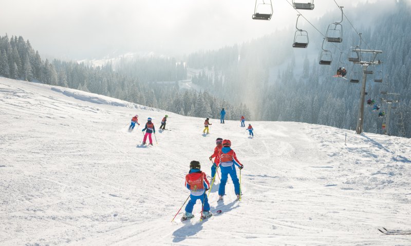 Kinder und Erwachsene fahren Ski auf den verschneiten Pisten eines Skigebiets in den Alpen an einem sonnigen Wintertag.