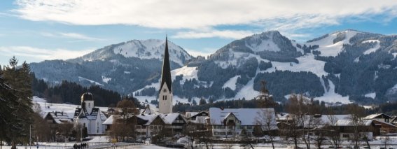 Das Dorf liegt in einer verschneiten Winterlandschaft, umgeben von majestätischen Bergen unter einem blauen Himmel.
