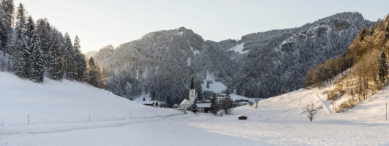 Eine schneebedeckte Landschaft mit einem Bauernhof, umgeben von Bergen und Bäumen während des Tages in den Alpen.