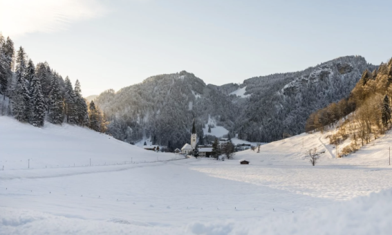 Eine schneebedeckte Landschaft mit einem Bauernhof, umgeben von Bergen und Bäumen während des Tages in den Alpen.