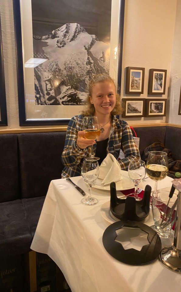 Eine junge Frau sitzt in einem eleganten Restaurant, sie hebt ein Glas und lächelt. Der Tisch ist festlich gedeckt.