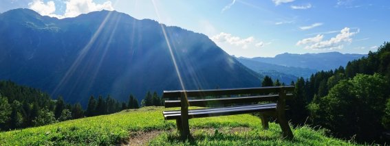 Eine Bank steht auf einer Wiese mit weitem Blick auf die Berge unter einem strahlend blauen Himmel.