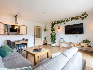 In einem hellen Raum stehen ein Sofa, ein Tisch und Pflanzen. Eine Wand hat einen großen Fernseher und eine gemütliche Atmosphäre.