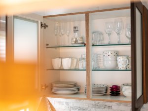 Ein Küchenschrank zeigt Geschirr und Gläser. Teller und Tassen stehen auf Regalen. Die Szene ist gut beleuchtet und ordentlich.