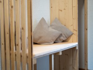 Eine Holzbank steht in einem Raum. Auf der Bank liegen zwei Kissen. Der Raum hat eine einfache Gestaltung.