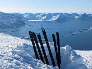 Mehrere Skier stecken im Schnee an einem Berg mit Blick auf fjordartige Landschaften und schneebedeckte Berge im Hintergrund.