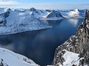 Schneebedeckte Berge umgeben einen tiefblauen Fjord an einem klaren Tag. Der Himmel ist blau und die Aussicht ist weit.