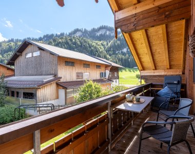 Sonniger Holzbalkon mit Tisch und Stühlen, Blick auf Nachbarhaus, Wiesen und bewaldete Berge – Bergheimat Ferienwohnungen in Obermaiselstein-Ried im Allgäu.
