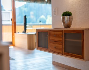 Heller Wohnraum mit Holz-Sideboard, Pflanze, Fernseher und bodentiefem Fenster – Bergheimat Ferienwohnungen in Obermaiselstein-Ried im Allgäu.
