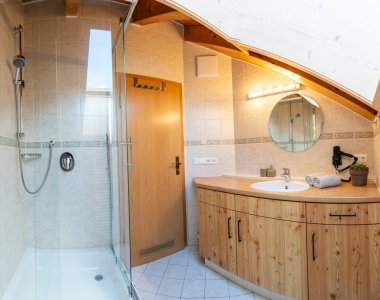 Helles Bad unter der Dachschräge mit Glasdusche, Waschtisch aus Holz, rundem Spiegel und Dachfenster – Bergheimat Ferienwohnungen in Obermaiselstein-Ried im Allgäu.