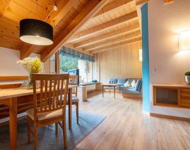 Wohnlicher Essbereich mit Holztisch und Blumen, dahinter Sitzecke und Fenster unter der Holzdecke – Bergheimat Ferienwohnungen in Obermaiselstein-Ried im Allgäu.