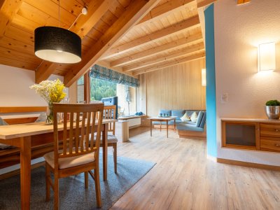 Wohnlicher Essbereich mit Holztisch und Blumen, dahinter Sitzecke und Fenster unter der Holzdecke – Bergheimat Ferienwohnungen in Obermaiselstein-Ried im Allgäu.