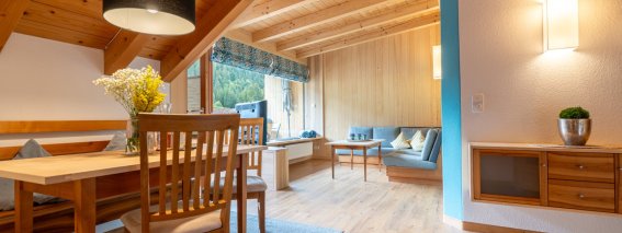 Wohnlicher Essbereich mit Holztisch und Blumen, dahinter Sitzecke und Fenster unter der Holzdecke – Bergheimat Ferienwohnungen in Obermaiselstein-Ried im Allgäu.
