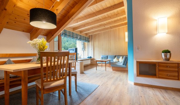 Wohnlicher Essbereich mit Holztisch und Blumen, dahinter Sitzecke und Fenster unter der Holzdecke – Bergheimat Ferienwohnungen in Obermaiselstein-Ried im Allgäu.