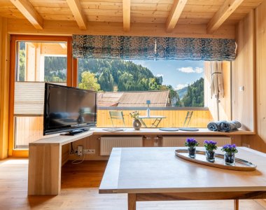 Wohnraum mit Holzmöbeln, Fernseher, Couchtisch und großem Fenster mit Blick auf Wald und Berge – Bergheimat Ferienwohnungen in Obermaiselstein-Ried im Allgäu.