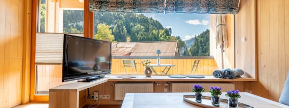Wohnraum mit Holzmöbeln, Fernseher, Couchtisch und großem Fenster mit Blick auf Wald und Berge – Bergheimat Ferienwohnungen in Obermaiselstein-Ried im Allgäu.