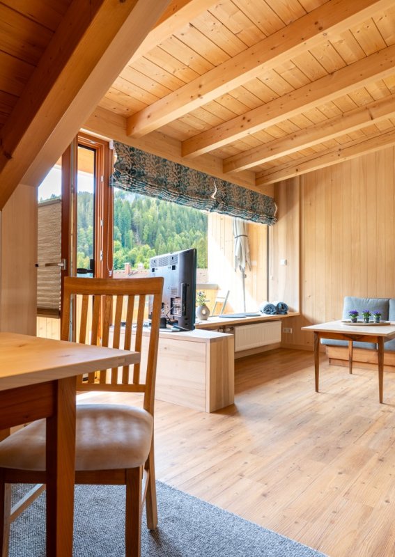 Offener Wohnbereich unter Holzbalken Großzügiger Wohn- und Essbereich unter sichtbaren Holzbalken mit Esstisch, Sitzecke und Fensterfront – Bergheimat Ferienwohnungen in Obermaiselstein-Ried im Allgäu.
