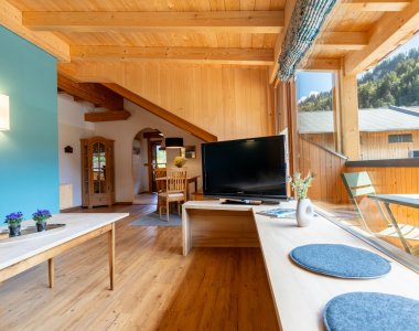 Heller Wohnraum mit Holzboden, TV, langer Fensterbank mit Sitzkissen und Blick auf die Berge – Bergheimat Ferienwohnungen in Obermaiselstein-Ried im Allgäu.