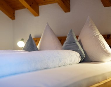 Doppelbett mit weißer Bettwäsche und blauen Kissen unter der Holzdecke – Bergheimat Ferienwohnungen in Obermaiselstein-Ried im Allgäu.