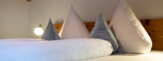 Doppelbett mit weißer Bettwäsche und blauen Kissen unter der Holzdecke – Bergheimat Ferienwohnungen in Obermaiselstein-Ried im Allgäu.