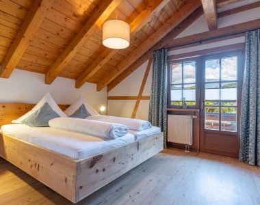Schlafzimmer unter Holzbalken mit massivem Doppelbett, Balkontür und Vorhängen – Bergheimat Ferienwohnungen in Obermaiselstein-Ried im Allgäu.