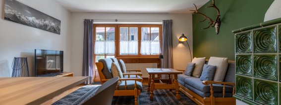 Großzügiges Wohnzimmer mit grünem Kachelofen, Sofa, Sesseln, Couchtisch und Hirschgeweih – Bergheimat Ferienwohnungen in Obermaiselstein-Ried im Allgäu.
