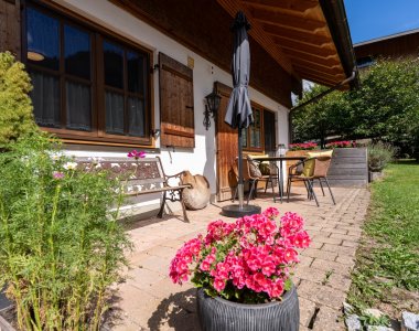 Terrasse mit pinken Blumen, Gartenbank, Sitzgruppe und Rasen vor der Ferienwohnung – Bergheimat Ferienwohnungen in Obermaiselstein-Ried im Allgäu.