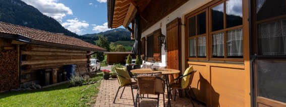 Gepflasterte Terrasse mit Tisch und Stühlen direkt vor der Wohnung, Blick auf Garten, Holzstadel und Berge – Bergheimat Ferienwohnungen in Obermaiselstein-Ried im Allgäu.