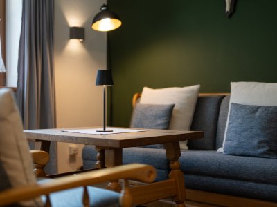Stilvolle Sitzecke mit Holztisch, Sessel, Sofa mit Kissen und Leuchten vor dunkelgrüner Wand – Bergheimat Ferienwohnungen in Obermaiselstein-Ried im Allgäu.